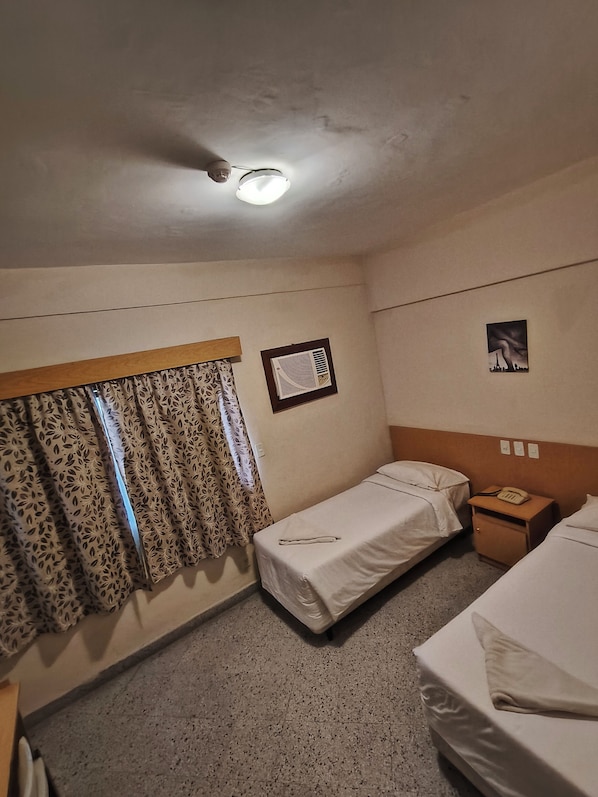 Habitación de hotel con dos camas individuales, una mesita de noche pequeña con teléfono, un radiador fijado a la pared y una ventana con cortinas estampadas.