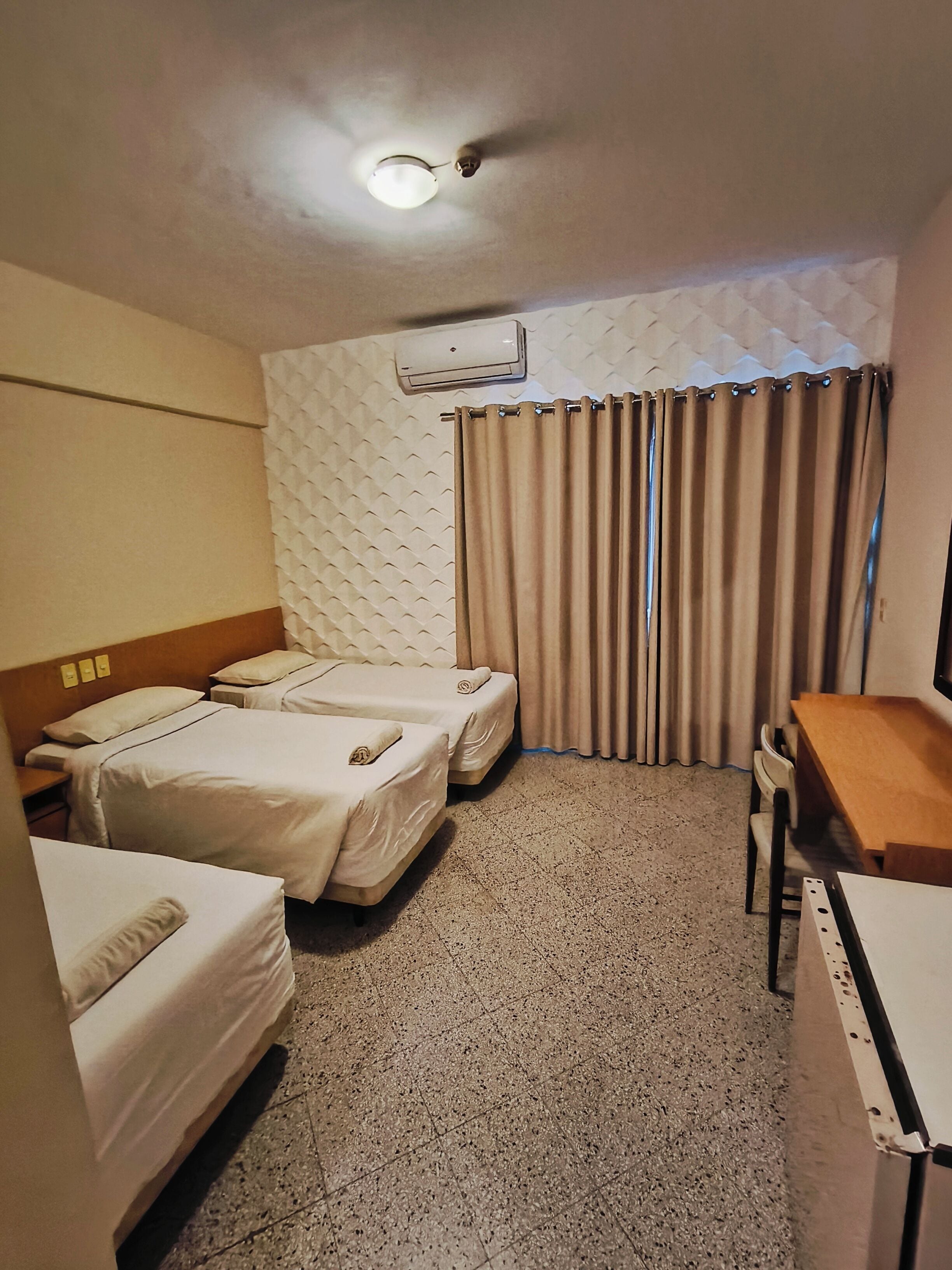 A hotel room with two beds, a desk, a chair, a ceiling fan, and a window with curtains.