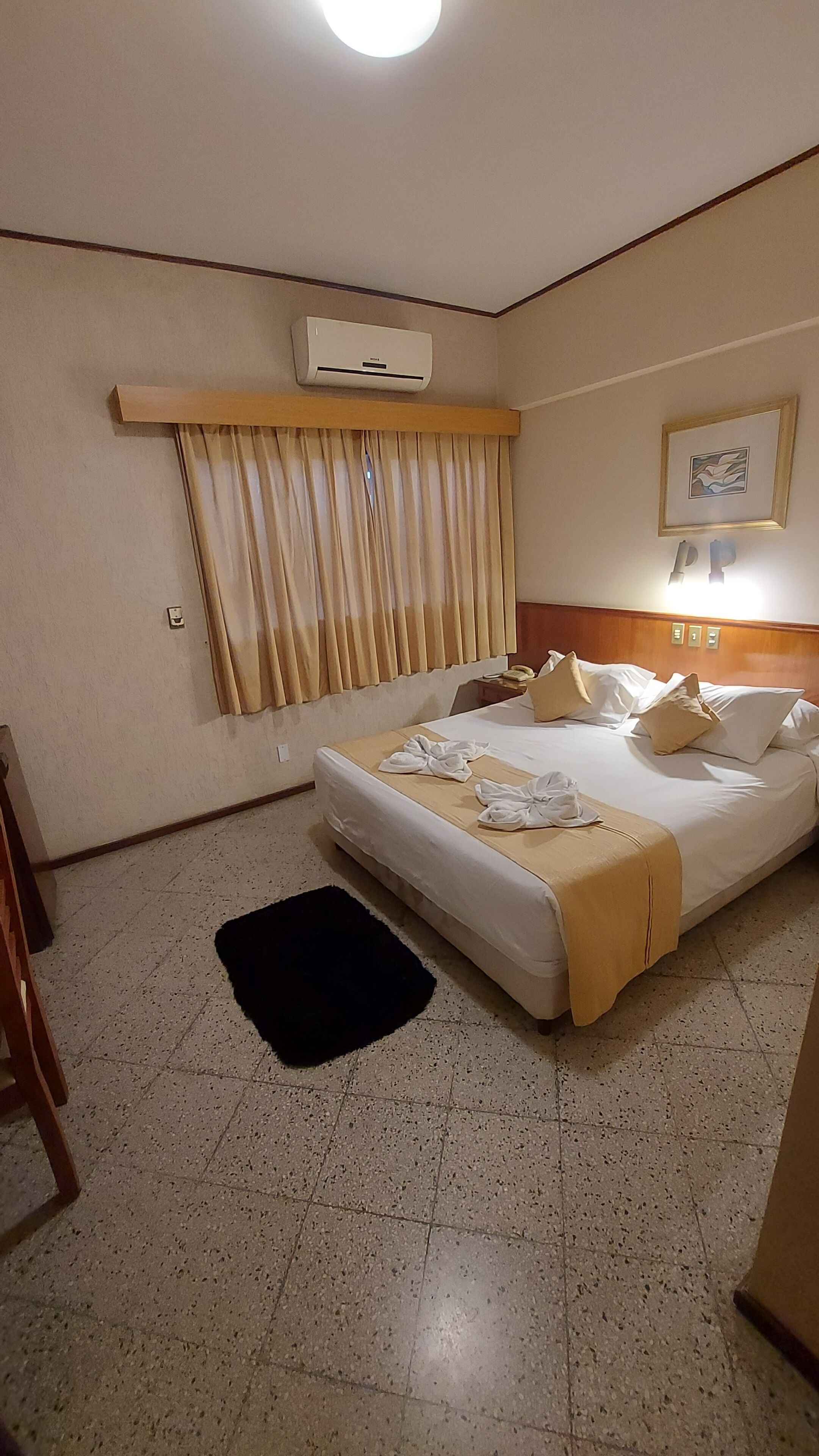 A hotel room with a bed, curtains, an air conditioner, a framed picture, and a telephone.