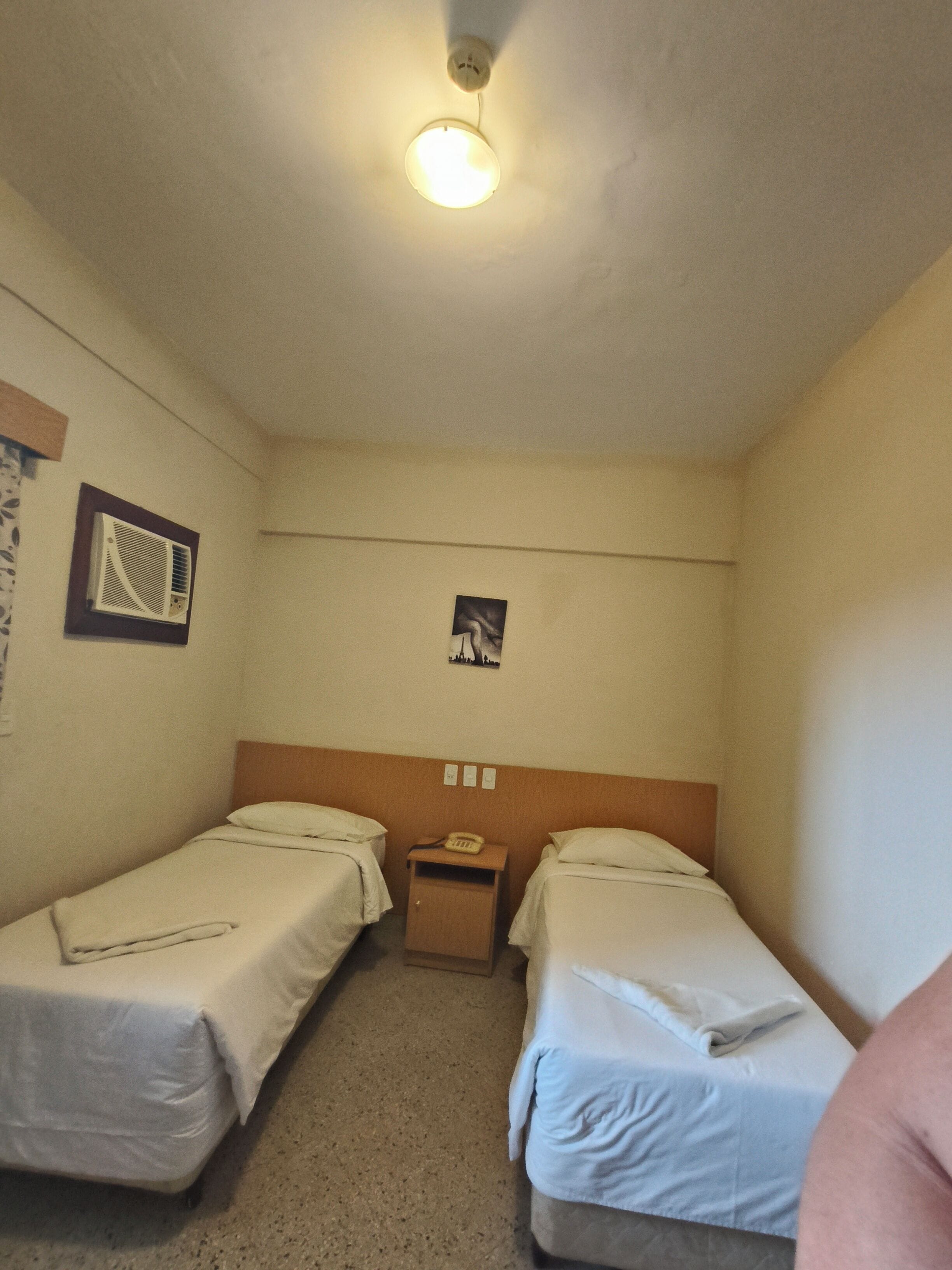 A small, single-room hotel with two single beds, a bedside table with a phone, a wall-mounted air conditioner, and a ceiling light.