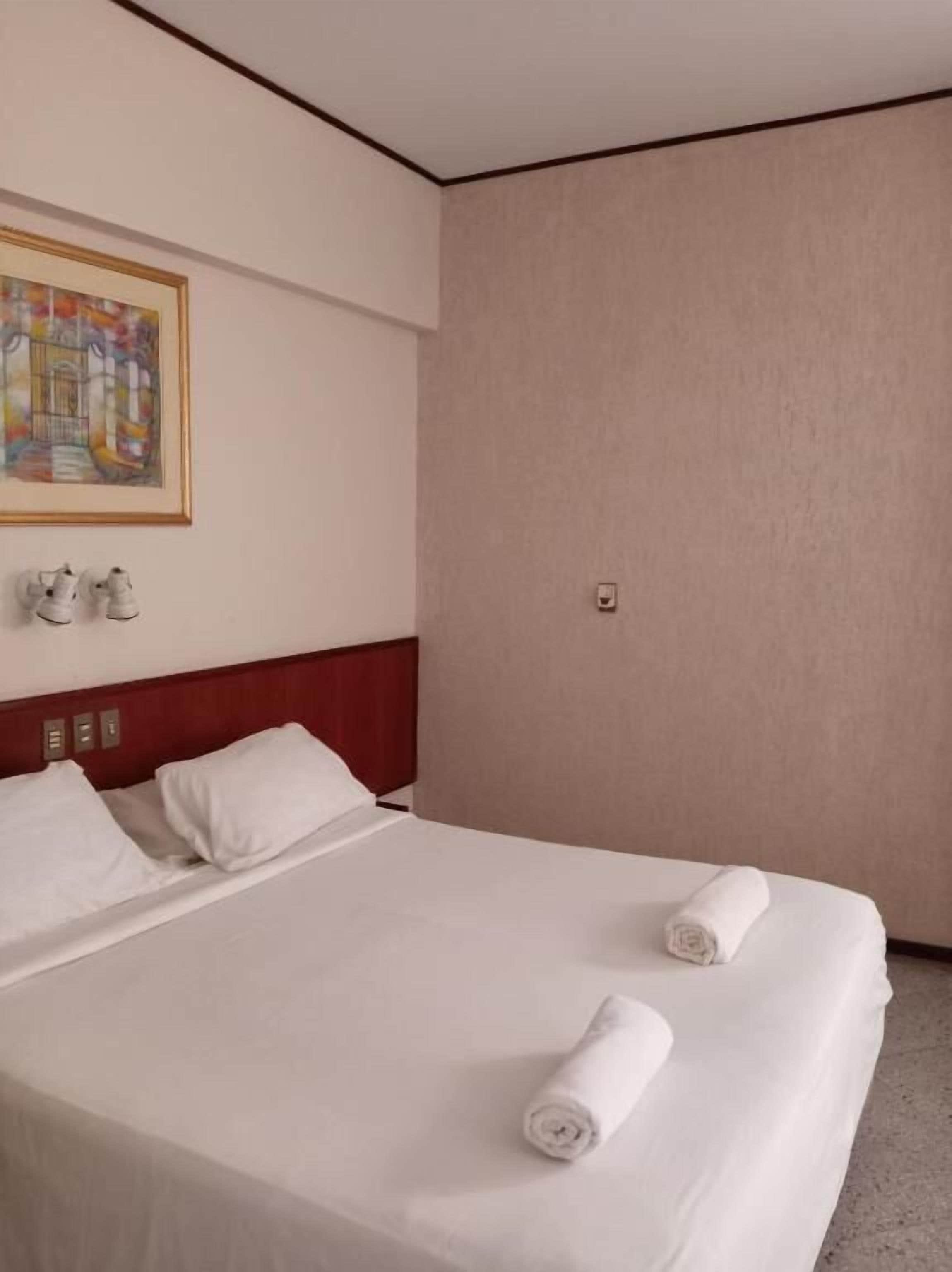 A hotel room with a bed, pillows, a headboard, a painting on the wall, and a pair of shoes on the floor.