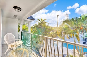 Un balcon avec une table et des chaises en verre, une piscine et des plantes tropicales.