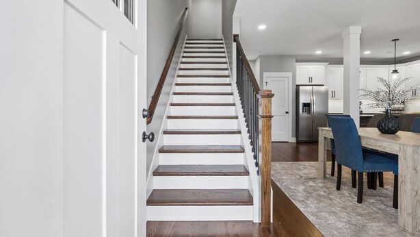 Un escalier mène à une cuisine dotée de meubles blancs, une table à manger avec des chaises et un réfrigérateur.