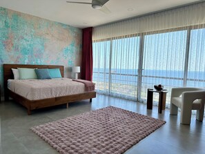 A bedroom with a large bed, a view of the sea, a ceiling fan, and a small table with a bowl.