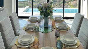 Een eetruimte met een tafel voor zes personen, grote ramen met uitzicht op de oceaan en heuvels, en een vaas met bloemen op tafel.