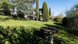 Un giardino curato con una panchina in pietra, alberi alti e un edificio sullo sfondo.
