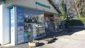 Una farmacia con un'ampia vetrina, un cartello con la scritta 'PHARMACIE' e un piccolo spazio esterno con vasi di piante.
