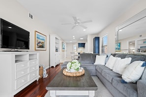A modern living room with a flat-screen TV, a ceiling fan, a grey sofa with white and blue cushions, and a coffee table with a floral arrangement.