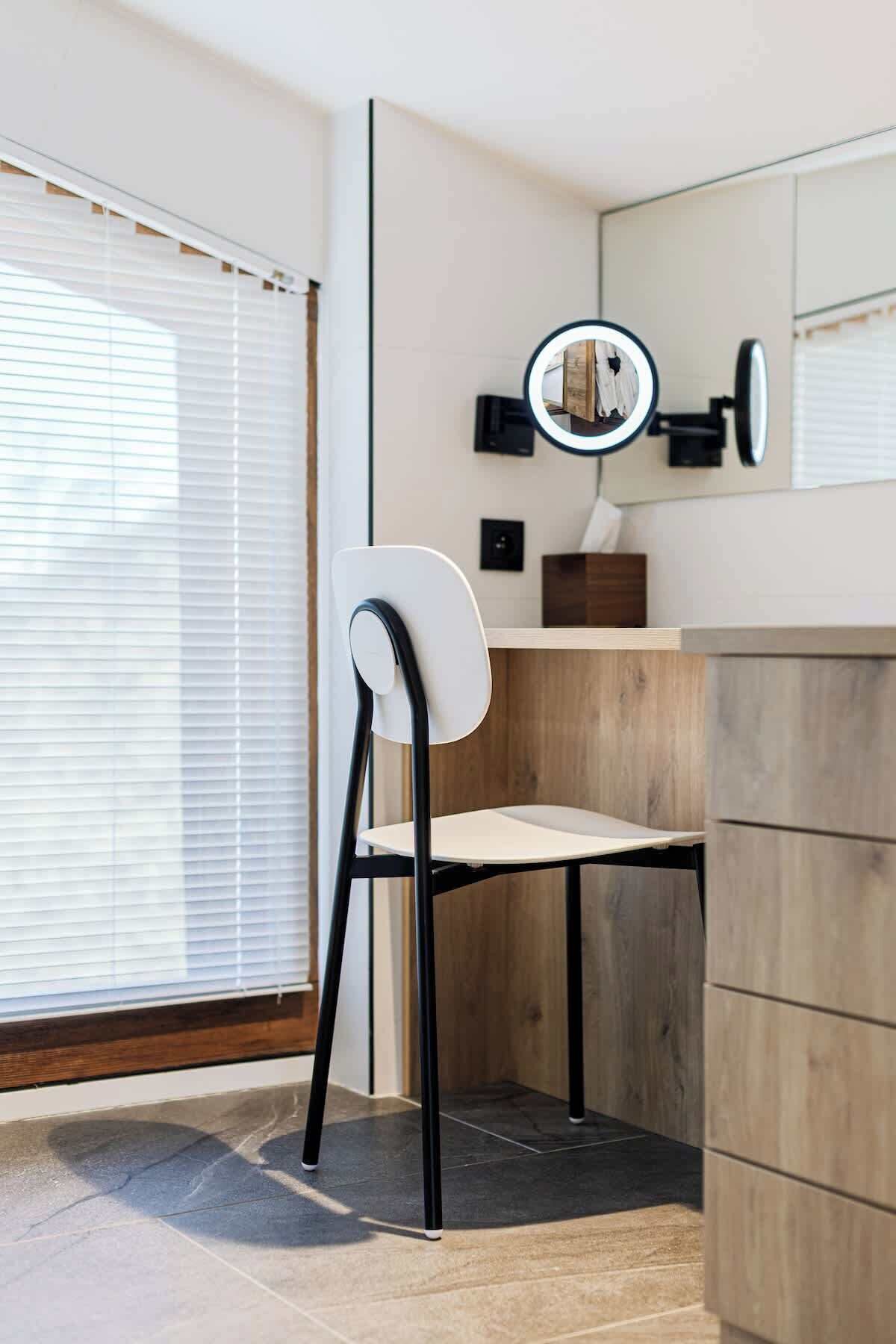 A modern bathroom with a white chair, a wooden vanity, a mirror with a ring light, and a window with blinds.