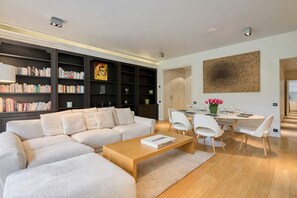 A modern living room with a sectional sofa, a wooden coffee table, a dining area with a white table and chairs, and a bookshelf filled with books. - Superb 2 bdr apartment Terrace Neuilly-sur-Seine (Neuilly-sur-Seine)