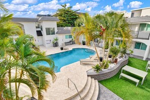 A swimming pool with a curved design, surrounded by palm trees and a grassy area with benches. - Stylish 1BR Suite w/ Pool & Steps to the Beach (Pompano Beach)