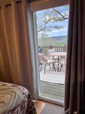 A bedroom with a large sliding glass door leading to an outdoor deck with wicker furniture and a view of trees and hills.