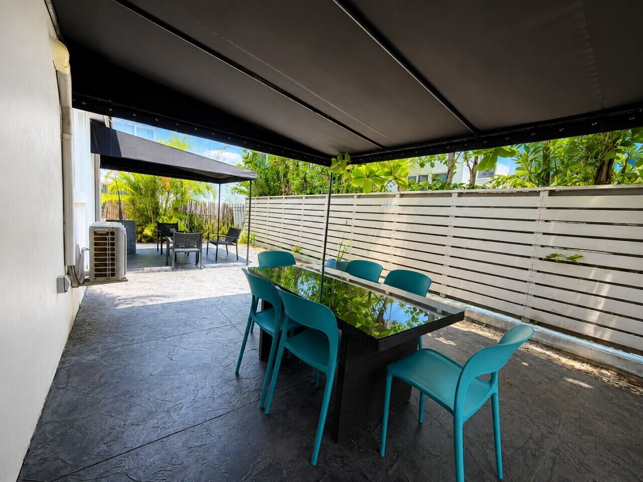 An outdoor patio with a table and chairs, a covered area, and a white fence in the background.