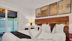 A hotel room with a large bed, a wooden headboard, a nightstand with a lamp, and a sliding glass door leading to an outdoor area. - Modern Beachside 2BR | Steps to Beach + Heated Pool (Pompano Beach)