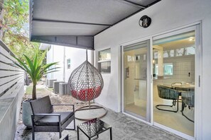 A covered patio area with outdoor furniture, a hanging chair, and a view into the house. - Coastal 1BR | Heated Pool | Steps to Beach (Pompano Beach)