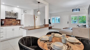 A modern kitchen with white cabinets, a wooden countertop, and a dining area with a round table set for four. - Modern 1BR Beachside | Heated Pool + Parking (Pompano Beach)