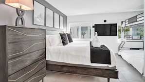 A modern hotel room with a large bed, a desk, a TV, and a balcony with plants. - Modern 1BR Beachside | Heated Pool + Parking (Pompano Beach)