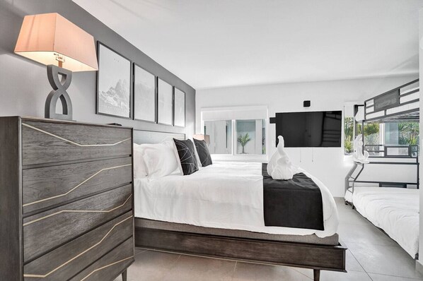 A modern hotel room with a large bed, a desk, a TV, and a balcony with plants. - Coastal 1BR | Heated Pool | Steps to Beach (Pompano Beach)