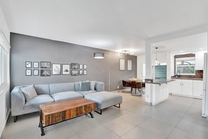 A modern living room with a grey sofa, a wooden coffee table, and a kitchen area with white cabinets and a window.
