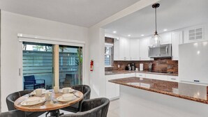 A modern kitchen with white cabinets, a granite countertop, and a dining area with a round table and chairs. - Modern 1BR Beachside | Heated Pool + Parking (Pompano Beach)
