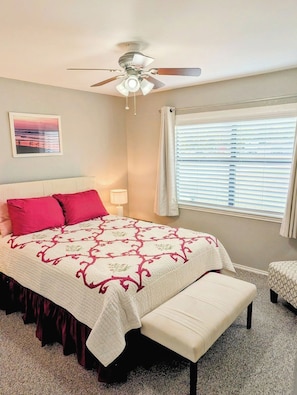 Une chambre à coucher comprenant un lit, une table de chevet, une lampe, un ventilateur de plafond, une fenêtre avec des stores et un tableau au mur.