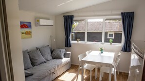 A room with a sofa, a dining table with chairs, a window with curtains, a wall-mounted air conditioner, and a small potted plant on the windowsill.