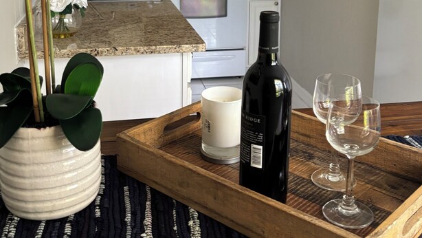Una cocina con armarios blancos, una estufa de acero inoxidable, una orquídea en maceta, una bandeja con una botella de vino y copas, y una taza blanca.