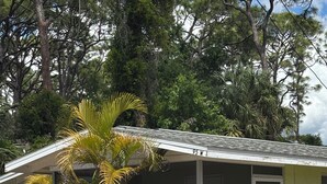 Un edificio de una planta con techo inclinado, cerca blanca en forma de piquete y una palmera al frente.