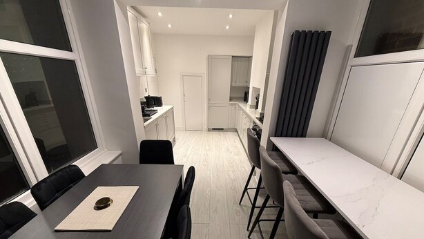 Et kompakt køkken med hvide skabe, en central ø og et spiseområde med et mørkt bord og stole.