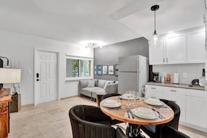 Une cuisine moderne avec des meubles blancs, un réfrigérateur encastré, une table à manger pour quatre personnes et un canapé gris dans l’espace salon.