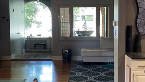 Una sala de estar con sofá, alfombra estampada, ventana con vistas al exterior y una zona de cocina al fondo.