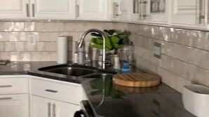 Una cocina con armarios blancos, un lavamanos de acero inoxidable, una tabla de cortar de madera y una planta.