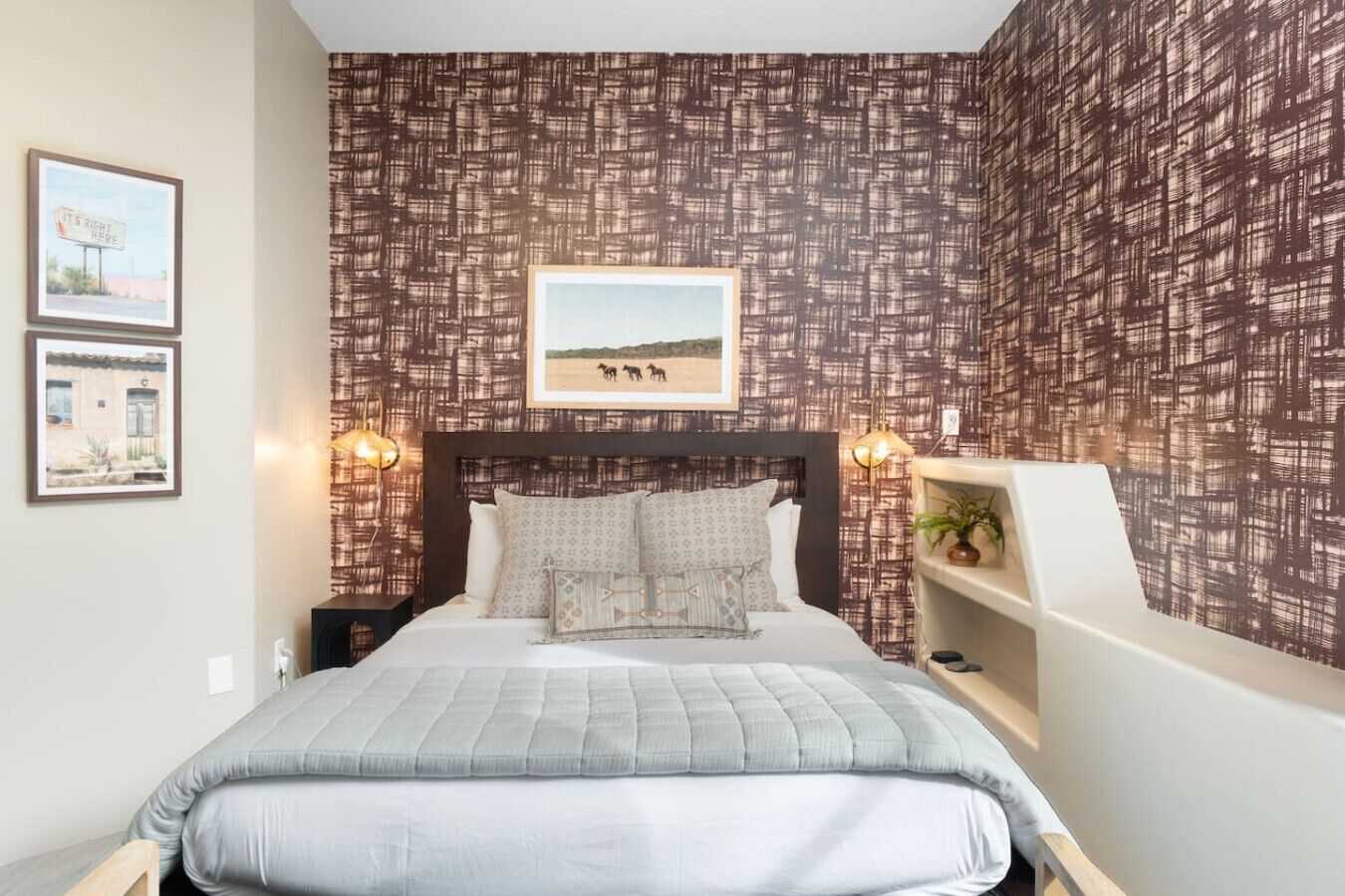 A bedroom with a patterned wallpaper, a bed with multiple pillows, a nightstand, a wall-mounted shelf, and two framed pictures.