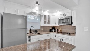 A modern kitchen with white cabinets, a granite countertop, a stainless steel refrigerator, a microwave, and a hanging light fixture. - Beach Across the Street | 1BR + Heated Pool (Pompano Beach)