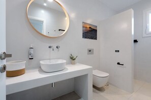 A modern bathroom with a round mirror, white sink, toilet, and a wall-mounted picture. - Villa Aura Kastraki NAXOS (Kastraki)