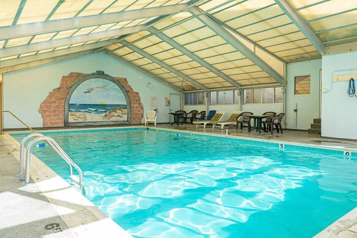 Ein beheiztes Hallenbad mit Fliesenboden, einer Metallleiter und einem Wandgemälde mit Strandmotiv.
