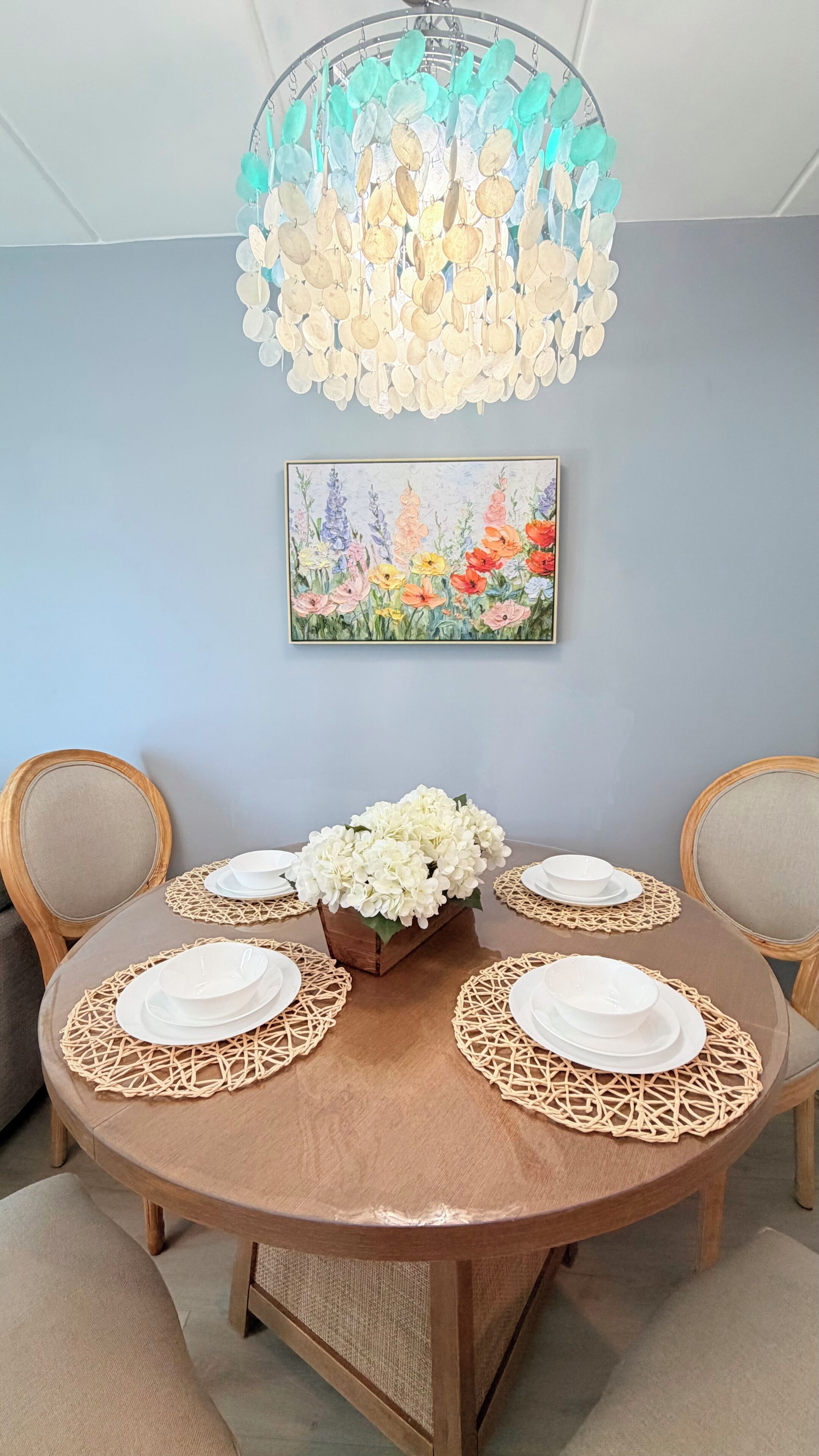 A dining table with a floral centerpiece, surrounded by chairs, with a decorative ceiling light and a floral painting on the wall.