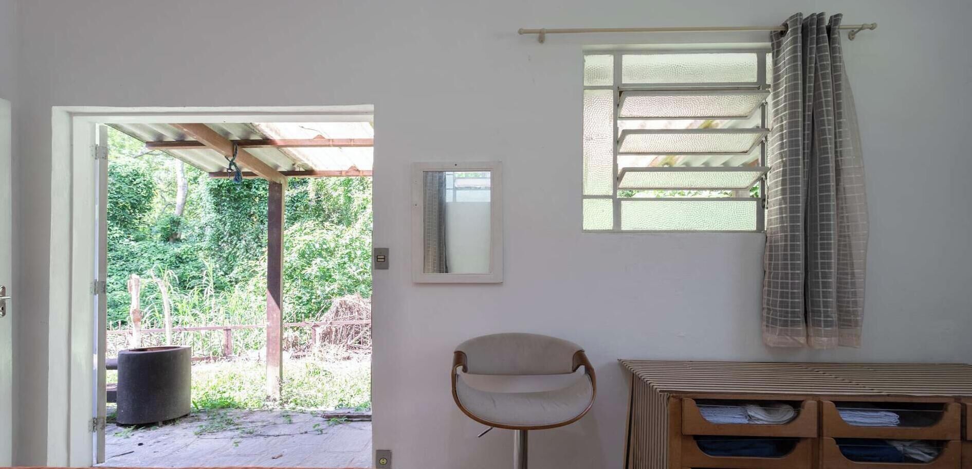 A room with a door leading to an outdoor area, a window with a curtain, a chair, and a small table with drawers.