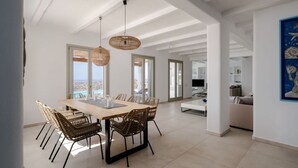 A spacious dining area with a wooden table and wicker chairs, large windows offering a view, and a modern kitchen in the background. - BLUE EYES VILLA Naxos Cyclades Greece (Naxos)