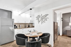 A modern kitchen with white cabinets, a grey refrigerator, and a dining area with a round table and black chairs. - 2BR Beach Escape | Heated Pool + Parking (Pompano Beach)