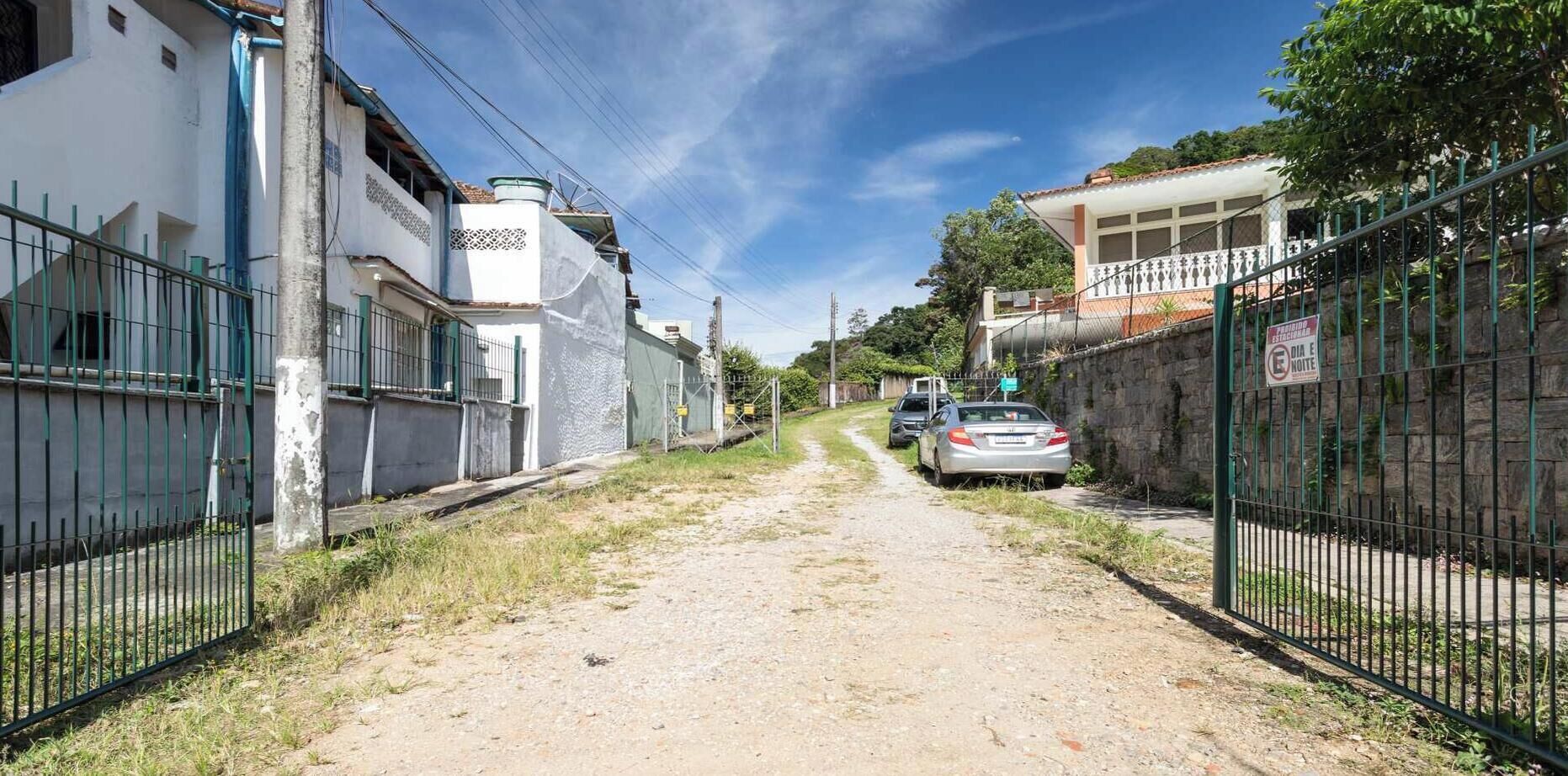 A narrow, unpaved alleyway flanked by buildings with metal fences, a parked car, and a clear sky.