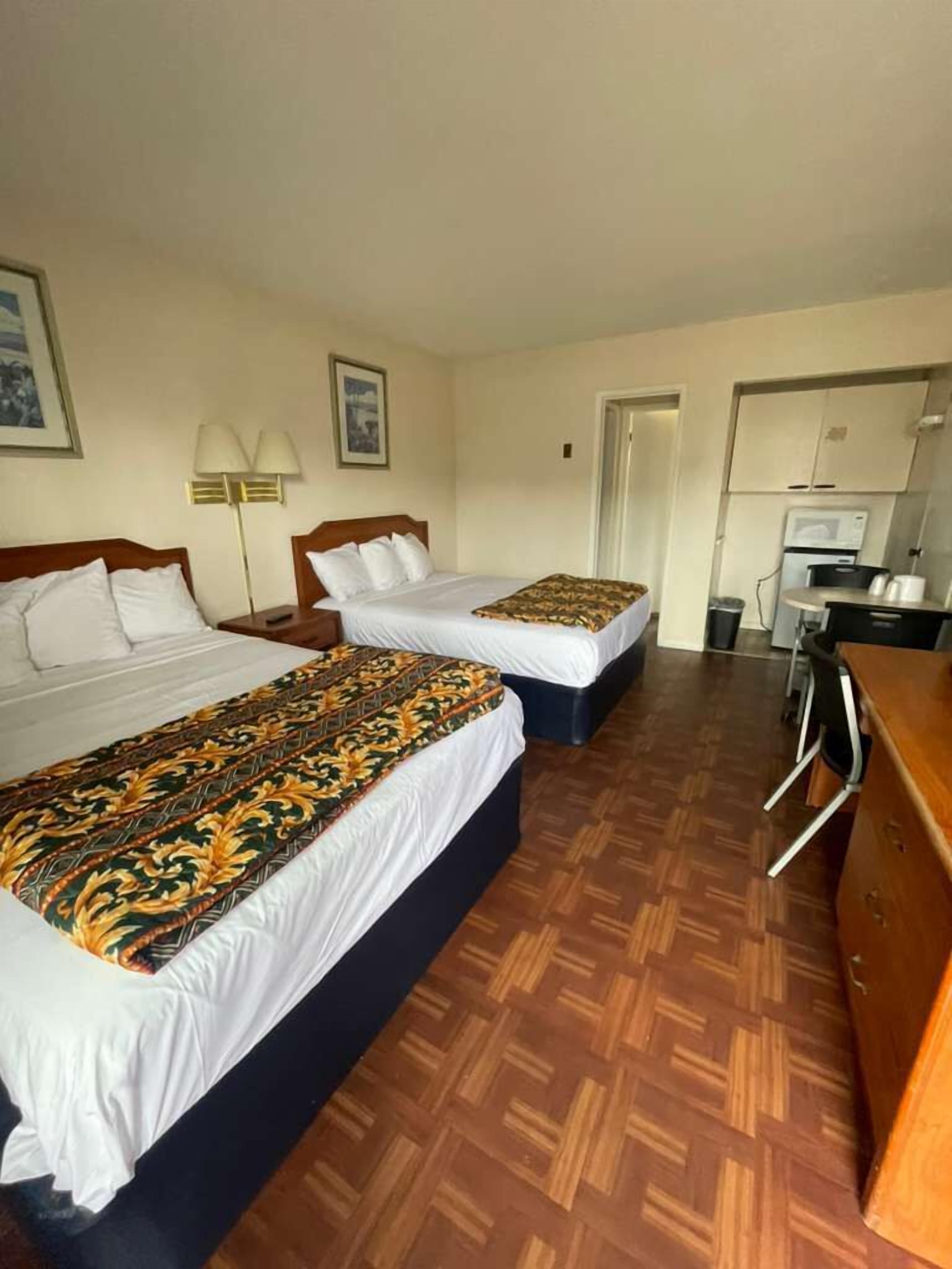 Habitación de hotel con dos camas, un escritorio, una silla, una lámpara y una kitchenette.