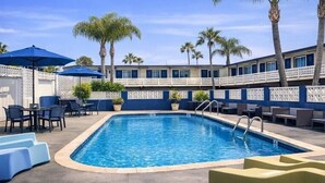 Una piscina con sillas de descanso y sombrillas, rodeada de palmeras y un edificio con balcones.