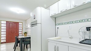 Una cocina con armarios blancos, un refrigerador, un lavamanos con un escurreplatos metálico y una zona de comedor con una mesa y sillas.