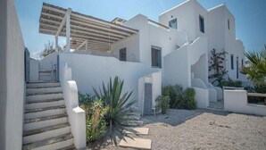 Un bâtiment moderne de couleur blanche, doté d’un escalier menant à une zone abritée, entouré de gravier et d’une végétation clairsemée.