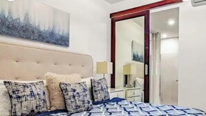 Un dormitorio con cabecera capitonada, ropa de cama estampada, una mesita de noche con lámpara y un espejo grande.