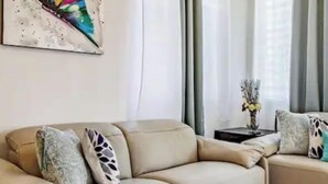 Una sala de estar con un sofá color beige, una mesita de noche con un florero, y un cuadro abstracto de colores en la pared.
