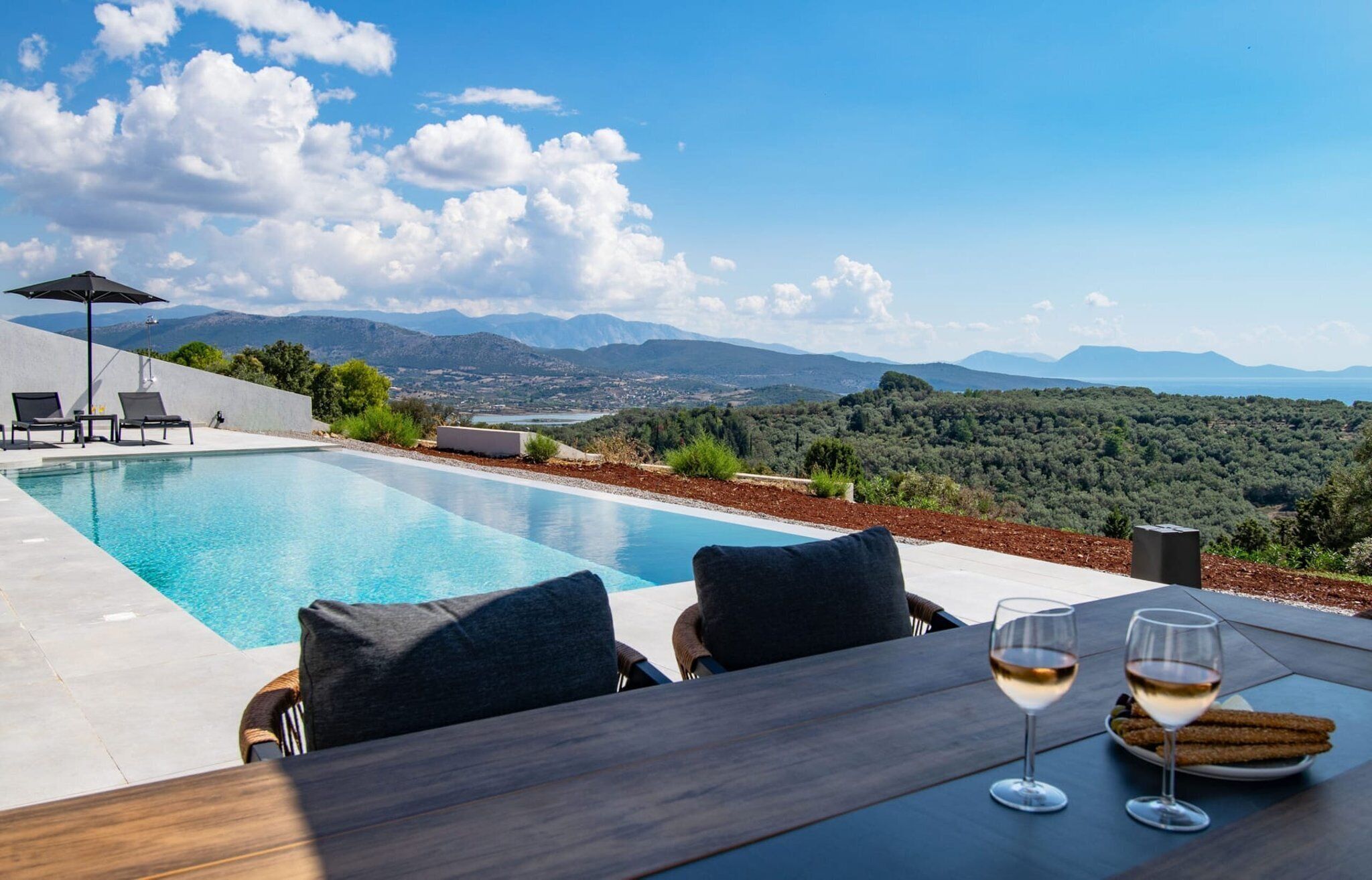 A pool with a view of mountains, two chairs, a table with glasses and snacks, and an umbrella.