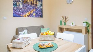 En matplats med ett bord dukat för två personer, en korg med frukt, en tallrik med bakverk, en klocka på väggen och en inramad tavla ovanför bordet.