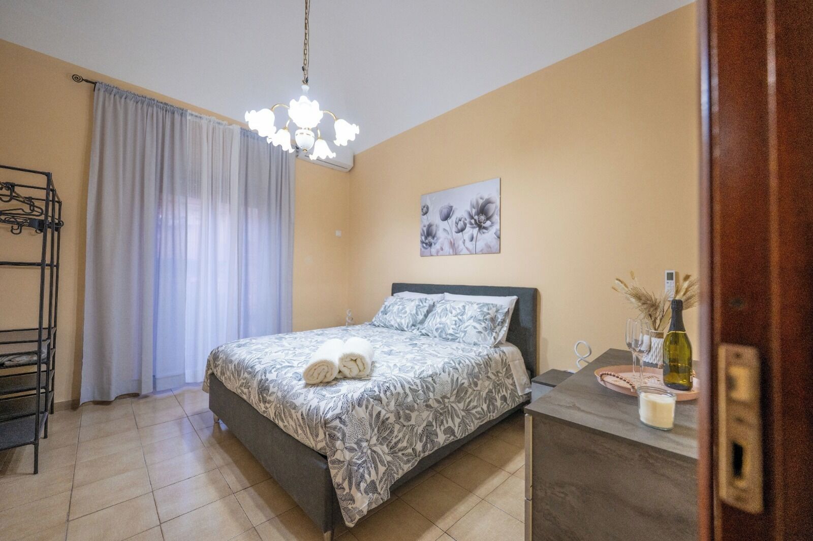 A bedroom with a bed, a chandelier, a painting, a bedside table with a bottle, and a window with curtains.
